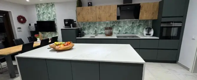 Küchenstudio Moderne Küche mit Kochinsel, grünen Schränken, Marmorrückwand und Holzakzenten. Esstisch und Stühle auf der linken Seite. Deckenleuchten erhellen den Raum.