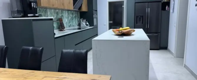 Küchenstudio Moderne Küche mit grauen Schränken, Marmorinsel und Geräten aus Edelstahl. Im Vordergrund steht ein Esstisch aus Holz mit schwarzen Stühlen. Auf der Insel steht eine Obstschale.