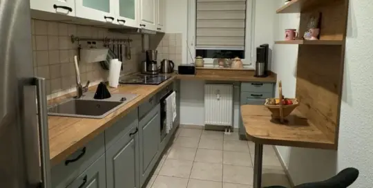 Küchenstudio Kompakte Küche mit grünen und weißen Schränken, Holzarbeitsplatten, Spüle, Herd und Geräten. Auf der rechten Seite befinden sich ein kleiner Tisch mit einem schwarzen Stuhl und Regale mit Dekor und einer Obstschale.