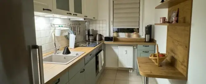 Küchenstudio Kompakte Küche mit weißen und grünen Schränken, Holzarbeitsplatten, Spüle, Ofen und kleinem Essbereich. Kaffeemaschine und Dekoartikel auf Regalen. Neutral gefliester Boden und Fenster mit Jalousie.