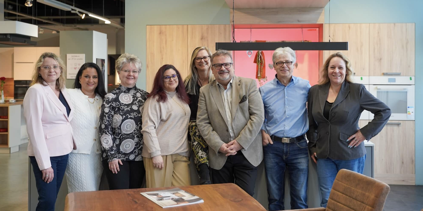 Küchenstudio Eine Gruppe von acht Personen steht zusammen in einem Küchenausstellungsraum und posiert für ein Foto. Sie tragen Business-Casual-Kleidung und lächeln. Zu sehen sind ein Holztisch und Küchenarmaturen.
