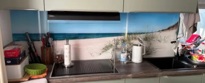 Küchenstudio Eine Küchentheke mit grünen Schränken, einer Spüle, Messern, einem Wasserkocher und einer Küchenrückwand im Stranddesign mit Sand und Meer.