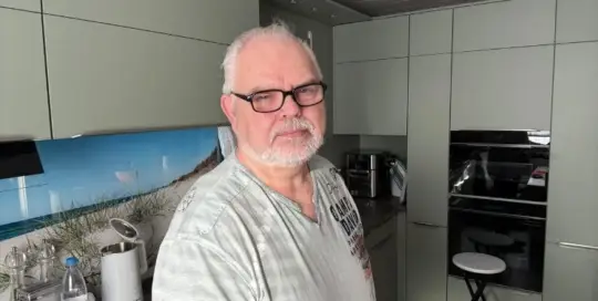Küchenstudio Ein Mann mit Brille und Bart steht in einer modernen Küche mit grauen Schränken und einem schwarzen Ofen.