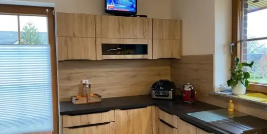 Küchenstudio Eine Küche mit Holzschränken, schwarzer Arbeitsplatte, einer Heißluftfritteuse, einer Kaffeemaschine und einem kleinen Fernseher darauf. Auf der rechten Seite befindet sich ein Fenster mit Jalousien und einer Topfpflanze.