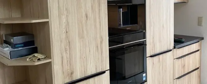 Küchenstudio Küchenschränke aus Holz mit eingebauten Öfen und schwarzen Griffen. Offene Regale auf der linken Seite bieten Platz für Kartons und Putzlappen. Weiß gefliester Boden.