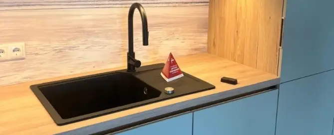 Küchenstudio Moderne Küche mit hellblauen Schränken, Holzarbeitsplatte und einer schwarzen Spüle mit gebogenem Wasserhahn. Neben der Spüle befindet sich ein kleines rotes Warnschild. Die Küchenrückwand zeigt eine ruhige Strandszene.