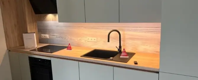 Küchenstudio Moderne Küche mit weißen Schränken, Holzarbeitsplatte, schwarzer Spüle und Herd. Unterschrankbeleuchtung erhellt den Bereich. Zwei kleine rote dreieckige Objekte und ein weißes Heft liegen bereit.