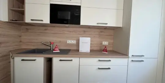 Küchenstudio Moderne Küche mit weißen Schränken, Holzarbeitsplatten, einer Spüle, eingebauter Mikrowelle und zwei kleinen roten Warnschildern neben dem Wasserhahn und auf der Arbeitsplatte.