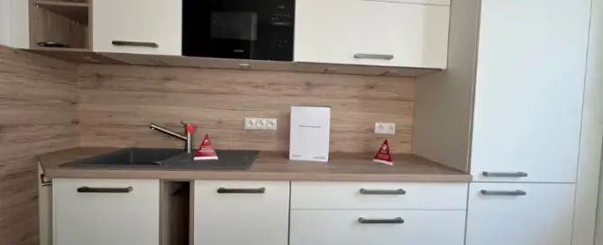 Küchenstudio Moderne Küche mit weißen Schränken, Holzarbeitsplatten, einer Spüle, eingebauter Mikrowelle und zwei kleinen roten Warnschildern neben dem Wasserhahn und auf der Arbeitsplatte.