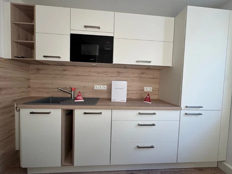 Küchenstudio Moderne Küche mit weißen Schränken, Holzarbeitsplatten, einer Spüle, eingebauter Mikrowelle und zwei kleinen roten Warnschildern neben dem Wasserhahn und auf der Arbeitsplatte.