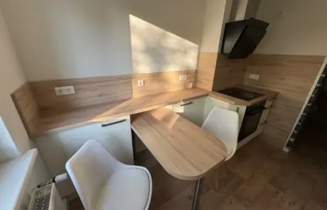 Küchenstudio Kompakte Küche mit Holzarbeitsplatten, zwei weißen Stühlen, einem Elektroherd mit Dunstabzugshaube und Einbauschränken. Sonnenlicht wirft Schatten auf Wand und Arbeitsplatte.