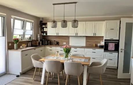 Küchenstudio Moderne Küche mit weißen Schränken, Holzakzenten, einem Esstisch mit sechs beigen Stühlen, Pendelleuchten und frischen Tulpen auf dem Tisch und der Theke.