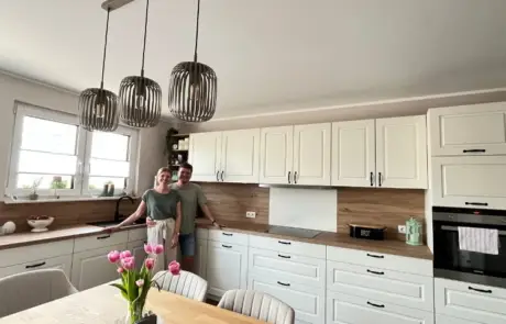 Küchenstudio Zwei Personen stehen zusammen in einer modernen Küche mit weißen Schränken, Holzarbeitsplatten und Hängelampen; auf dem Tisch im Vordergrund steht eine Vase mit rosa Tulpen.