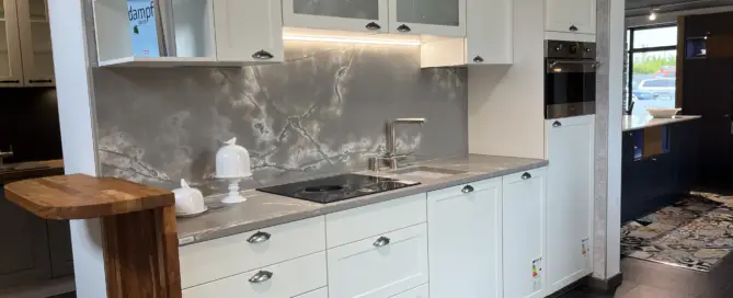 Küchenstudio Modernes Küchendisplay mit weißen Schränken, Einbaubackofen, Kochfeld, Metallspüle, Unterschrankbeleuchtung und Holzarbeitsplattenerweiterung auf der linken Seite - jetzt in unseren Ausstellungsküchen abverkaufbar.
