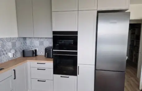 Küchenstudio Moderne Küche mit hellgrauen Schränken, einem Kühlschrank aus Edelstahl, einem eingebauten Backofen, einer Arbeitsplatte aus Holz, einer Heißluftfritteuse und einem Toaster vor einer gemusterten Aufkantung.
