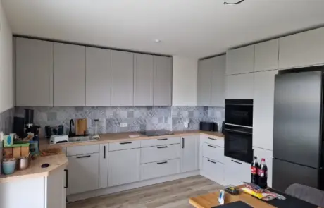 Küchenstudio Moderne L-förmige Küche mit hellgrauen Schränken, Holzarbeitsplatten, Einbaugeräten, einem schwarzen Kühlschrank und verschiedenen Gegenständen auf der Theke und dem Esstisch.