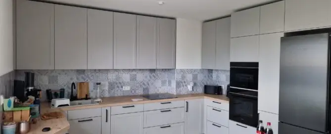 Küchenstudio Moderne L-förmige Küche mit hellgrauen Schränken, Holzarbeitsplatten, Einbaugeräten, einem schwarzen Kühlschrank und verschiedenen Gegenständen auf der Theke und dem Esstisch.