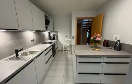 Küchenstudio Moderne Küche mit hellgrauen Schränken, Einbaugeräten, einer Spüle und einer Arbeitsplatte, die mit einer Blumenvase, einem roten Wasserkocher und einer Kaffeemaschine dekoriert ist.