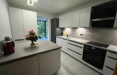 Küchenstudio Moderne Küche mit weißen Schränken, grauen Arbeitsplatten, einem Elektroherd, einer Spüle, schwarzen Geräten, einem roten Wasserkocher und einer Vase mit rosa Blumen auf der Insel; großes Fenster am anderen Ende.