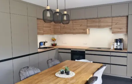 Küchenstudio Moderne Küche mit beigen und hölzernen Schränken, Einbaugeräten, einem Esstisch aus Holz mit Stühlen, Pendelleuchten und einer kleinen Pflanze als Mittelpunkt.