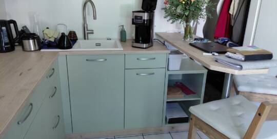 Küchenstudio Moderne Küche mit hellgrünen Schränken, einer Kaffeemaschine, einer Blumenvase auf einem Tresen, einem Barhocker und verschiedenen Küchen- und Büroartikeln auf den Arbeitsflächen.