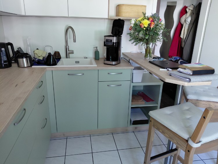 Küchenstudio Moderne Küche mit hellgrünen Schränken, einer Kaffeemaschine, einer Blumenvase auf einem Tresen, einem Barhocker und verschiedenen Küchen- und Büroartikeln auf den Arbeitsflächen.