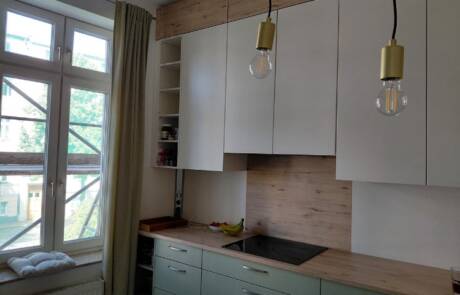 Küchenstudio Moderne Küche mit hellen Holz- und weißen Schränken, grünen Schubladen, eingebautem Herd, offenen Regalen und zwei Hängelampen, mit einem Fenster und Lichtvorhang auf der linken Seite.