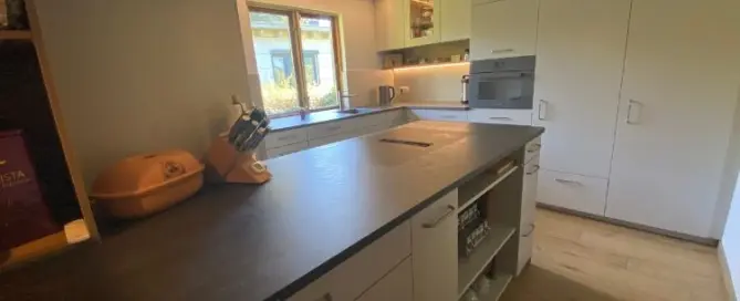 Küchenstudio Moderne Küche mit weißen Schränken, schwarzer Arbeitsplatte, eingebautem Ofen, Spüle unter einem Fenster und verschiedenen Küchenutensilien auf der Arbeitsplatte. Durch das Fenster fällt natürliches Licht ein.