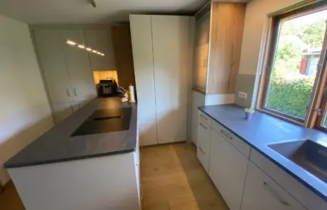 Küchenstudio Moderne Küche mit weißen Schränken, schwarzer Arbeitsplatte, Insel mit eingebautem Kochfeld, Holzfußboden, Spüle am Fenster und Kaffeemaschine in der Ecke.