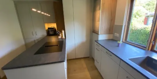 Küchenstudio Moderne Küche mit weißen Schränken, schwarzer Arbeitsplatte, Insel mit eingebautem Kochfeld, Holzfußboden, Spüle am Fenster und Kaffeemaschine in der Ecke.