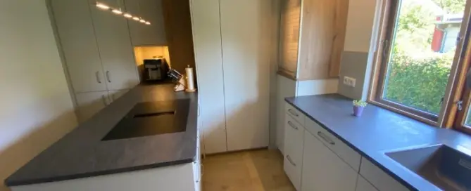 Küchenstudio Moderne Küche mit weißen Schränken, schwarzer Arbeitsplatte, Insel mit eingebautem Kochfeld, Holzfußboden, Spüle am Fenster und Kaffeemaschine in der Ecke.