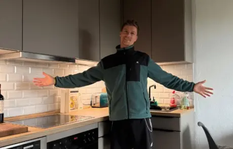 Küchenstudio Eine Person steht in einer modernen Küche mit ausgestreckten Armen und lächelt. Die Küche ist mit grauen Schränken, weißen Fliesen und verschiedenen Kleingeräten auf dem Tresen ausgestattet.