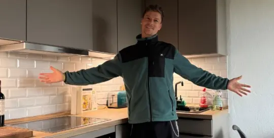 Küchenstudio Eine Person steht in einer modernen Küche mit ausgestreckten Armen und lächelt. Die Küche ist mit grauen Schränken, weißen Fliesen und verschiedenen Kleingeräten auf dem Tresen ausgestattet.