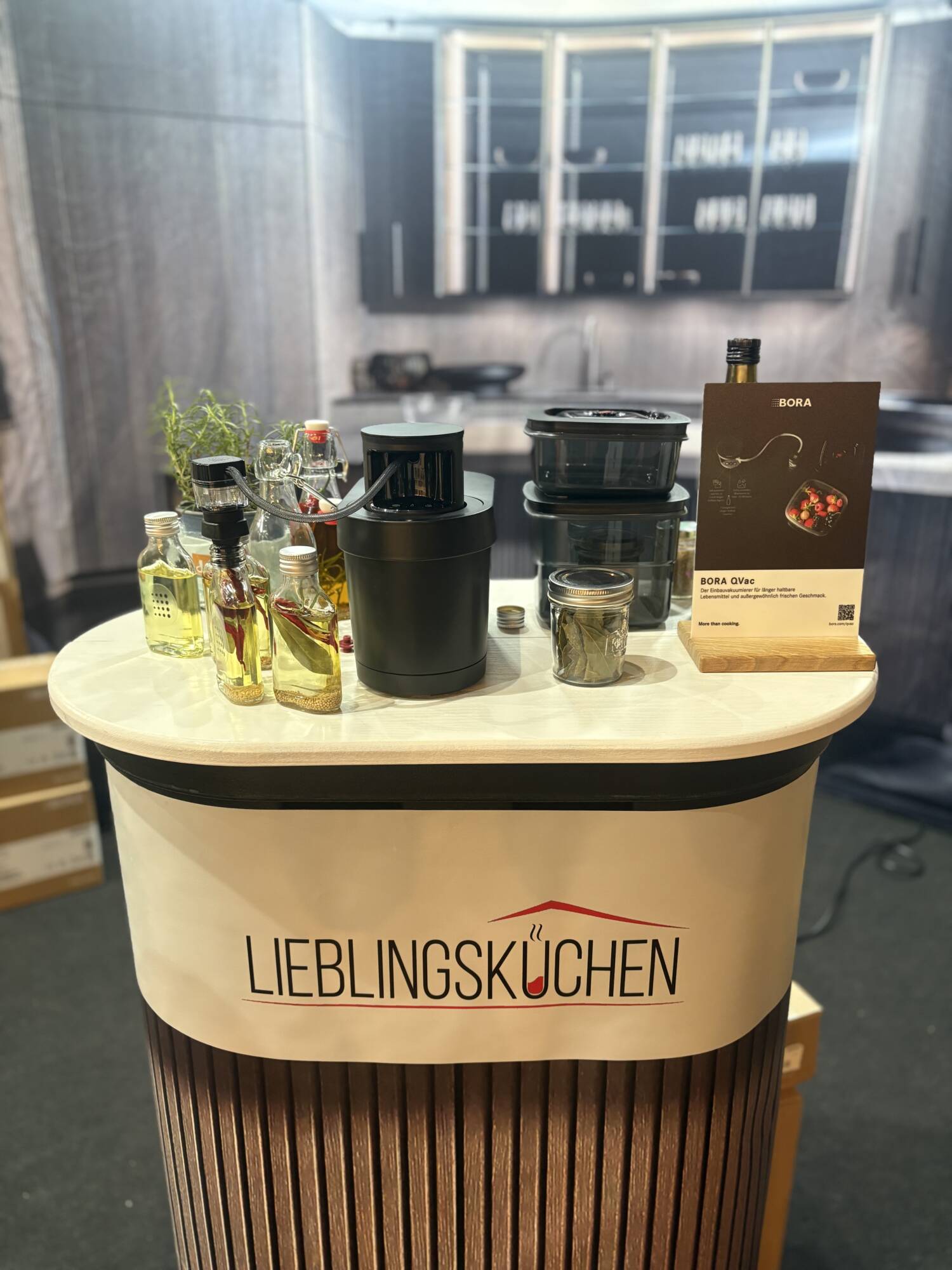 Küchenstudio Eine Küchenauslage mit Ölgläsern, Kräutern, Flaschen, Küchenutensilien und einer Produktbroschüre auf einem Ständer mit der Aufschrift "Lieblingsküchen" vor einem modernen Küchenhintergrund.