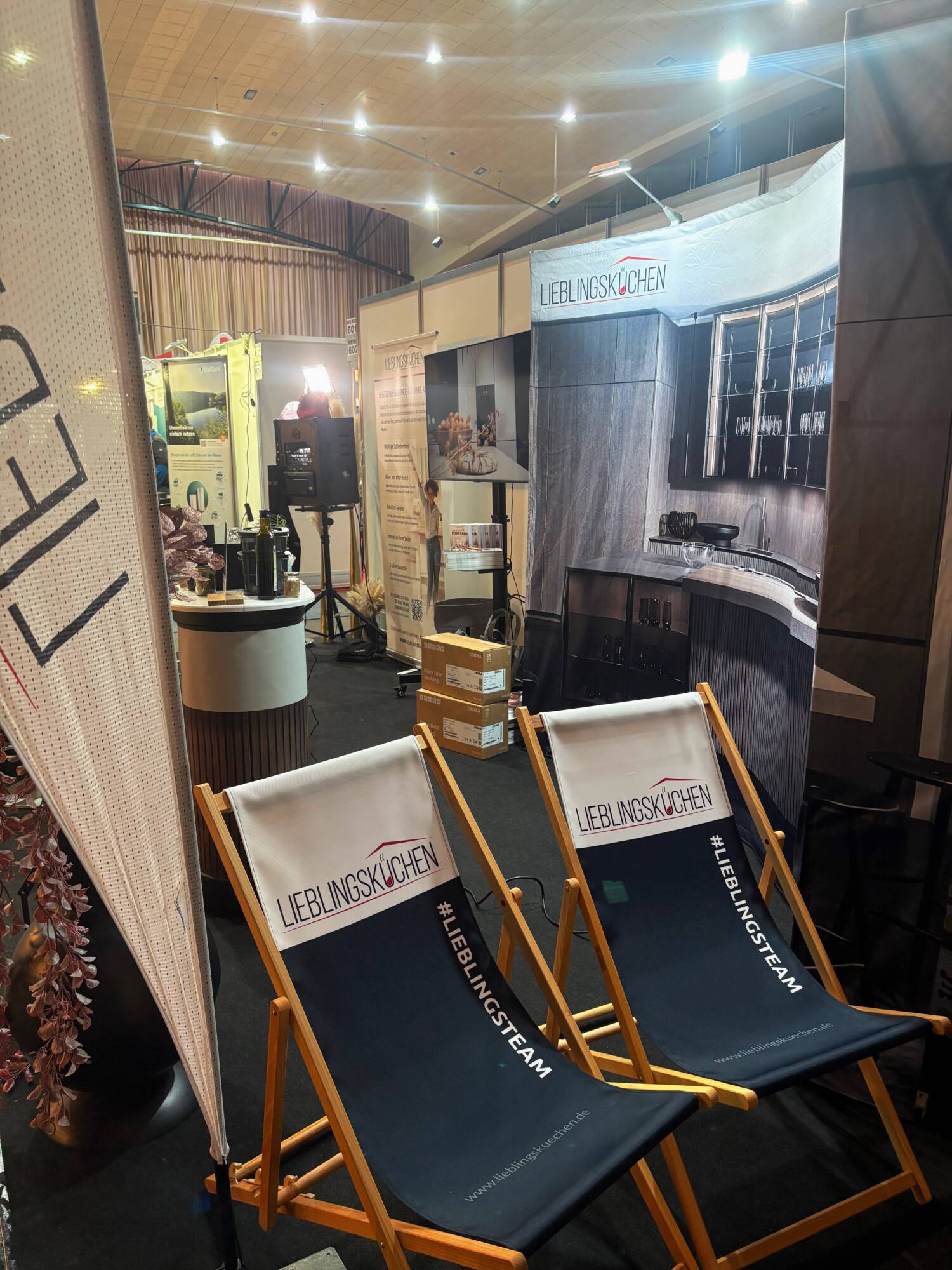 Küchenstudio Zwei Marken-Liegestühle und ein großer Küchenständer an einem überdachten Messestand, im Hintergrund sind Boxen und Beleuchtungsanlagen zu sehen.