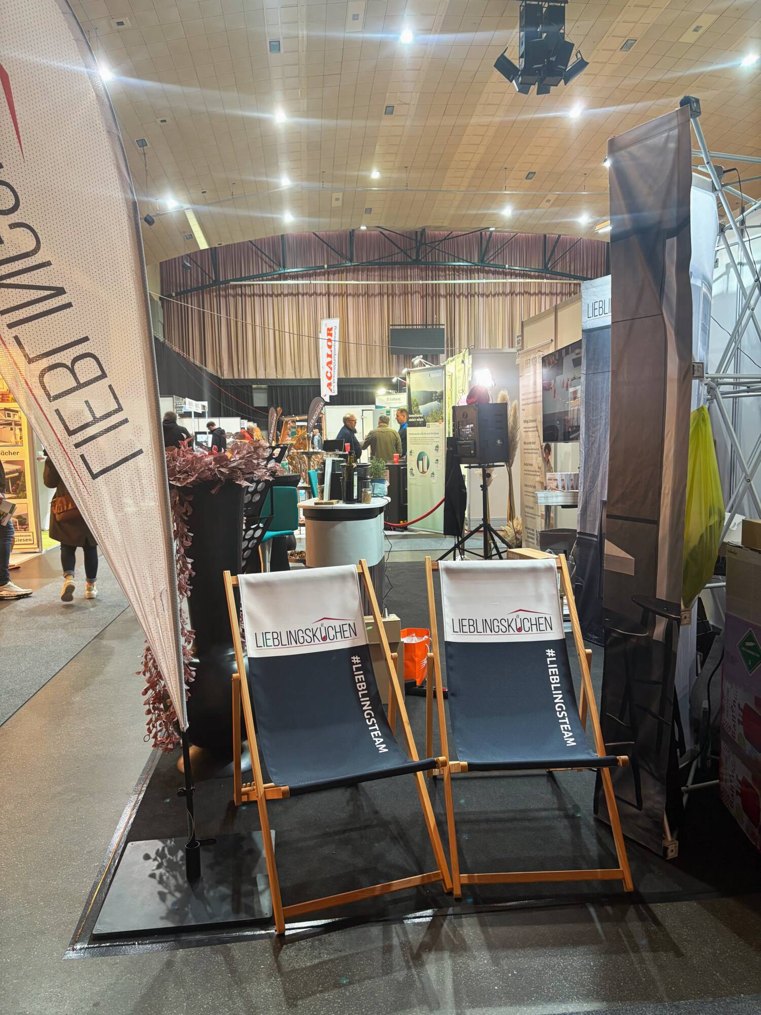 Küchenstudio Zwei Liegestühle mit dem Aufdruck "LIEBLINGSKÜCHEN" stehen an einem Messestand in einer großen überdachten Ausstellungsfläche. Im Hintergrund sind andere Stände und Besucher zu sehen.