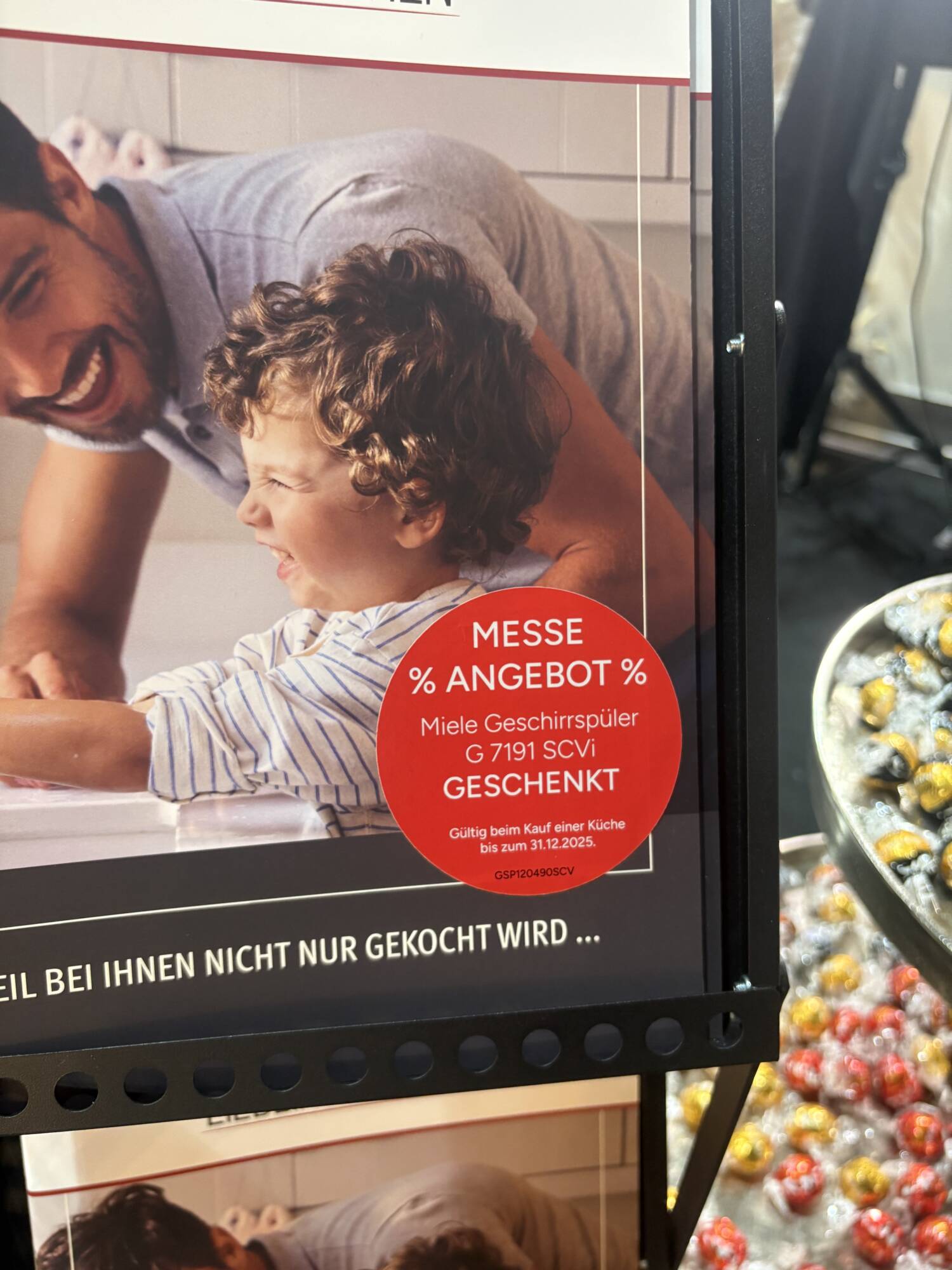 Küchenstudio Ein roter Werbeaufkleber auf einem Display bietet einen kostenlosen Miele-Geschirrspüler beim Kauf einer Küche an, gültig bis zum 31. Dezember 2025. Im Hintergrund sind ein Mann und ein Kind zu sehen.