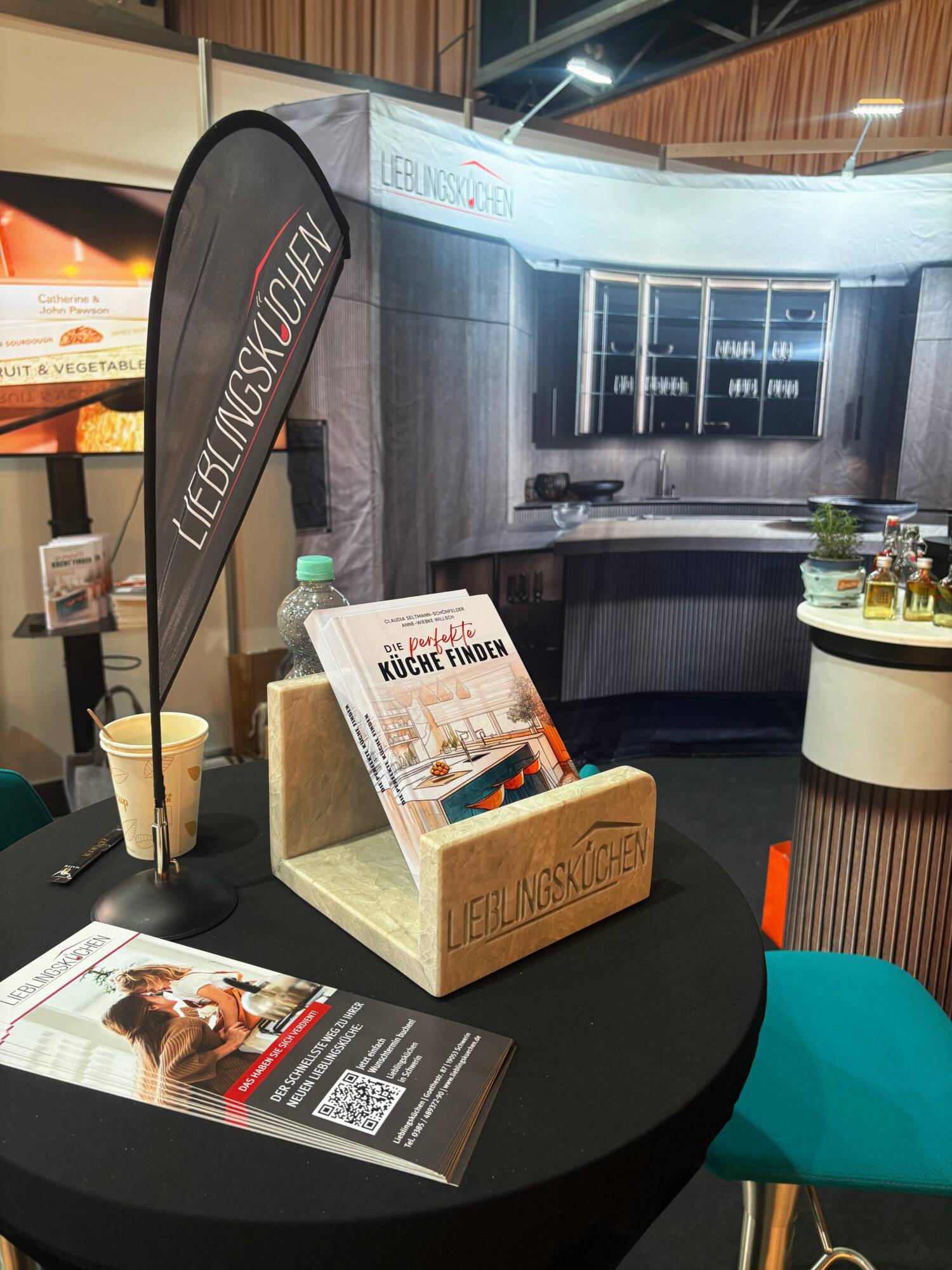 Küchenstudio Ein Messestand von LIEBLINGSKÜCHEN zeigt Broschüren, eine Wasserflasche und eine Kaffeetasse vor einer modernen Küchenkulisse.