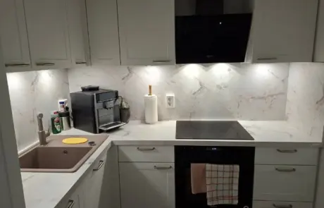 Küchenstudio Moderne Küche mit weißen Schränken und Marmorsockel, mit Spüle, Kaffeemaschine, Papierhandtuchhalter, Elektroherd, Backofen und Unterschrankbeleuchtung.