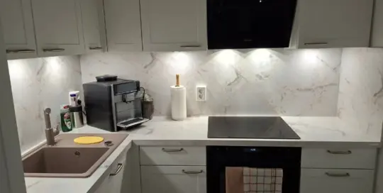 Küchenstudio Moderne Küche mit weißen Schränken und Marmorsockel, mit Spüle, Kaffeemaschine, Papierhandtuchhalter, Elektroherd, Backofen und Unterschrankbeleuchtung.