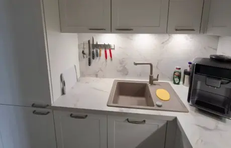 Küchenstudio Eine moderne Küchenspüle mit beigem Wasserhahn, Schneidebrett, Messern auf einer Magnetleiste, Spülmittel und einer Kaffeemaschine auf einer weißen Marmorarbeitsplatte.