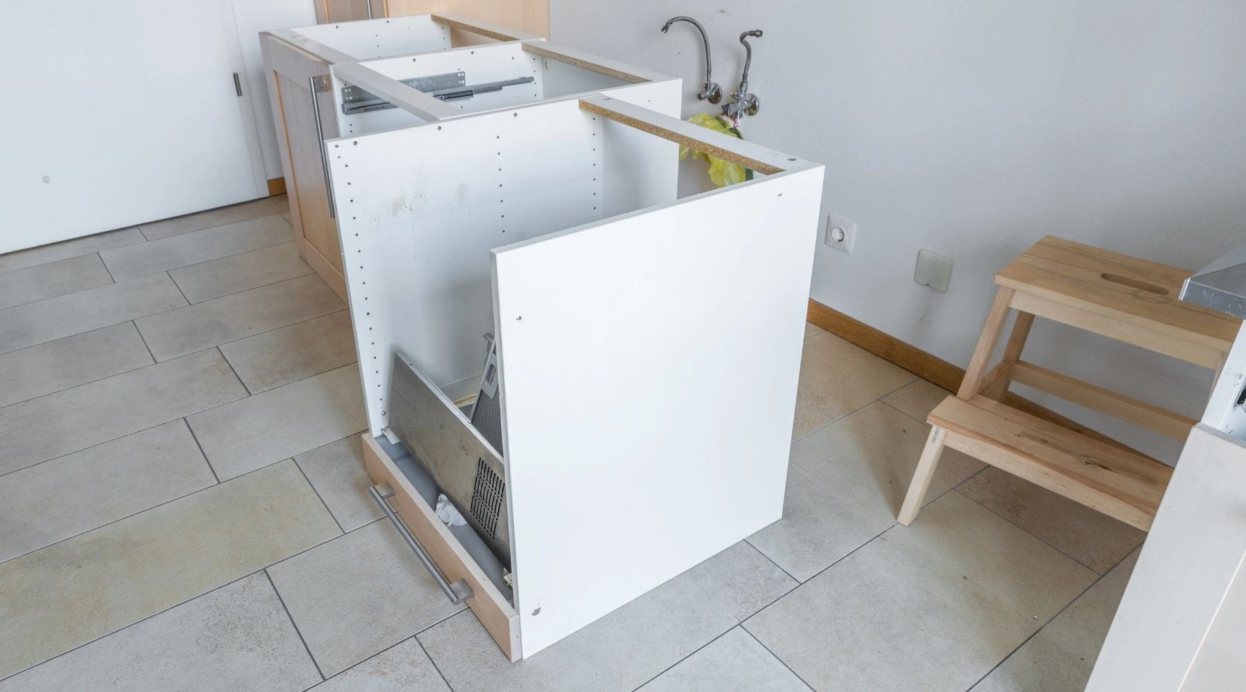 Küchenstudio Ein teilweise montierter weißer Küchenschrank mit Schublade steht auf einem gefliesten Boden; im Hintergrund sind Sanitärleitungen und ein hölzerner Tritthocker zu sehen.