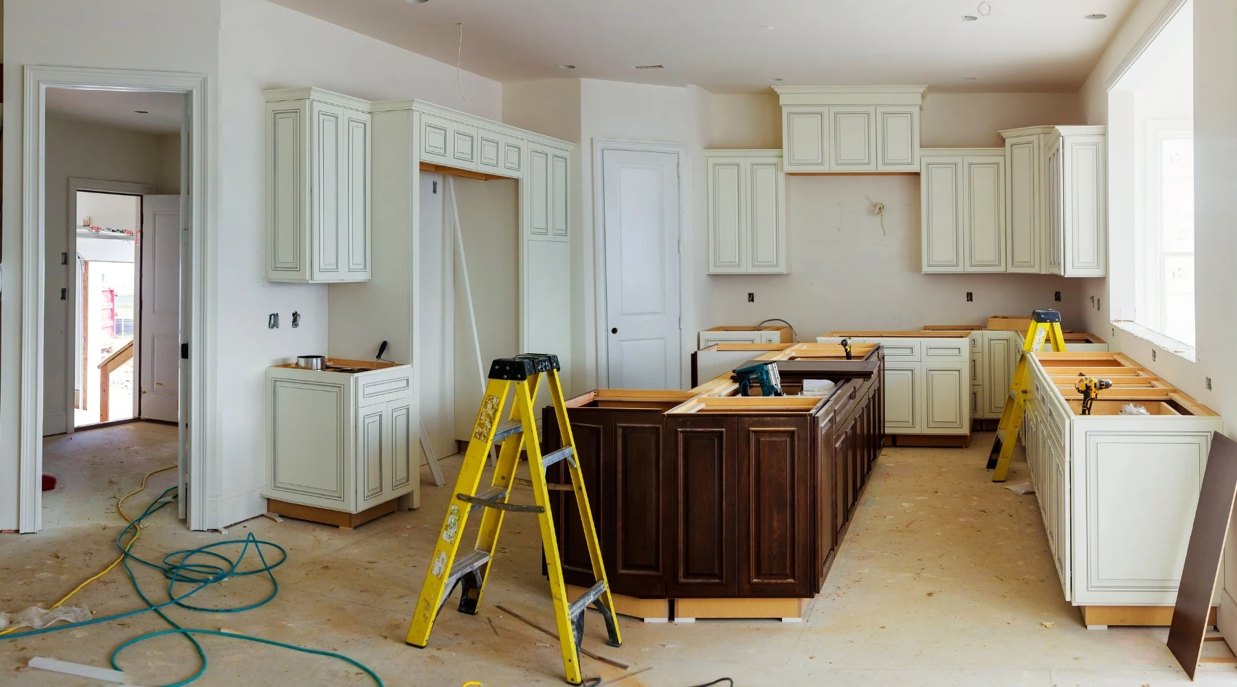 Küchenstudio Eine Küche im Bau mit weißen Schränken, einer dunklen Insel, Leitern, Werkzeugen und freiliegenden Wänden. Der Fußboden ist unfertig, und überall liegen Kabel und Materialien verstreut.