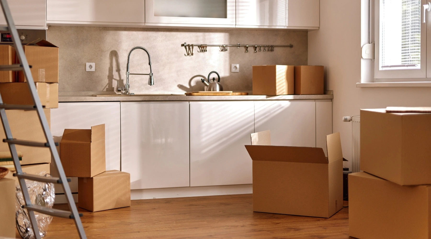 Küchenstudio Eine Küche mit weißen Schränken, einer Spüle und mehreren Umzugskartons, die auf dem Holzboden verstreut sind, bei natürlichem Tageslicht.