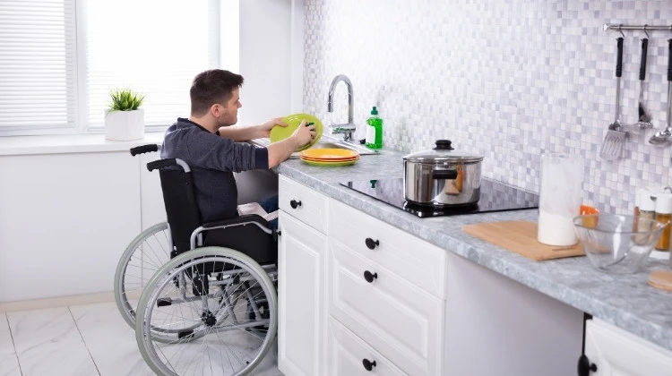Küchenstudio Eine Person im Rollstuhl wäscht das Geschirr an einer Spüle, während ein Topf auf dem Herd steht und verschiedene Utensilien an der Wand hängen.