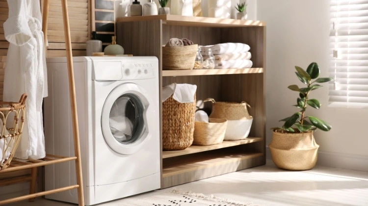 Küchenstudio In einer hellen, gut beleuchteten Waschküche steht eine Frontlader-Waschmaschine neben Holzregalen mit Körben, gefalteten Handtüchern und einer Topfpflanze.