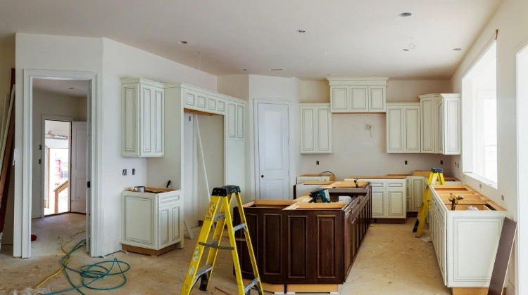 Küchenstudio Renovierte Küche mit weißen Schränken, einer zentralen braunen Insel, Werkzeugen, Leitern und unbearbeiteten Oberflächen. Helles natürliches Licht fällt durch große Fenster ein.