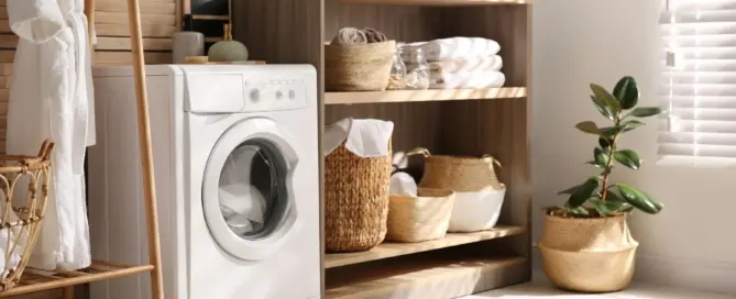 Küchenstudio Eine moderne Waschküche mit einer Frontlader-Waschmaschine, Holzregalen mit Körben, Handtüchern und Topfpflanzen, die durch Jalousien mit natürlichem Sonnenlicht erhellt werden.