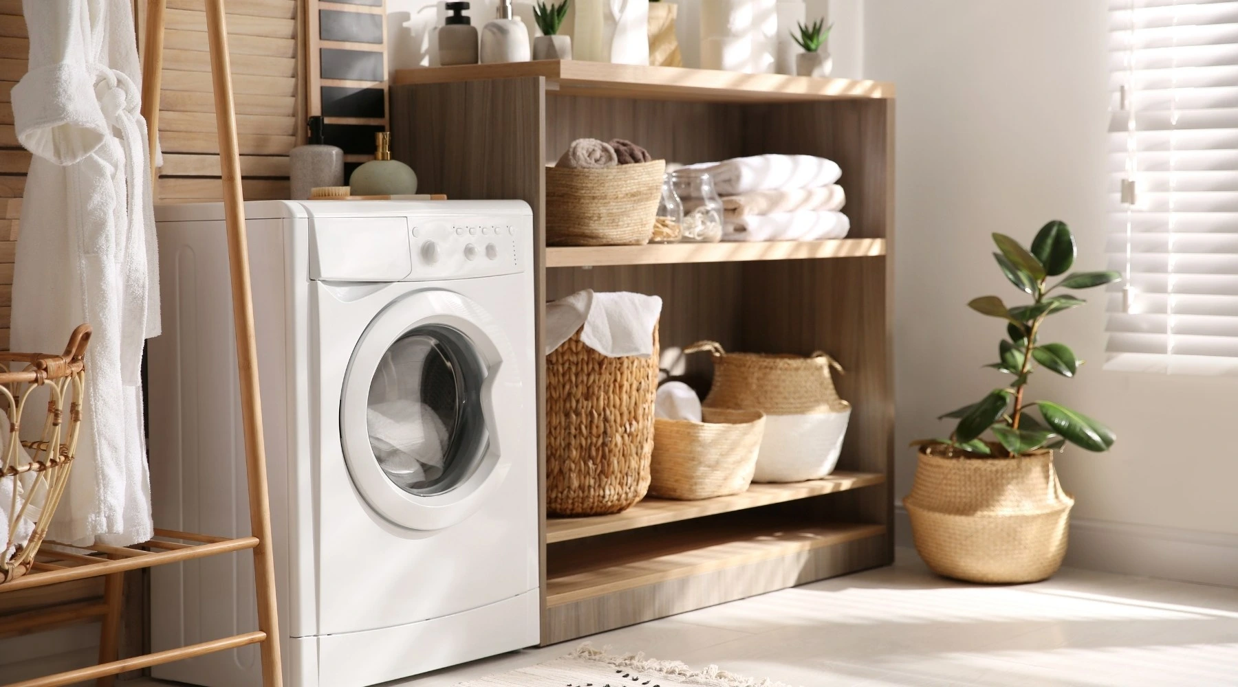 Küchenstudio Eine moderne Waschküche mit einer Frontlader-Waschmaschine, Holzregalen mit Körben, Handtüchern und Topfpflanzen, die durch Jalousien mit natürlichem Sonnenlicht erhellt werden.