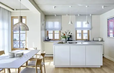 Küchenstudio Moderne Küche mit zentraler Insel, hellem Holzfußboden, Esstisch mit Stühlen, großen Fenstern und minimalistischem weißen Dekor.